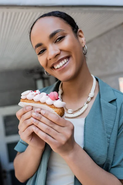 Fröhliche afrikanisch-amerikanische Frau mit köstlichem Eclair und Blick in die Kamera im Freien — Stockfoto