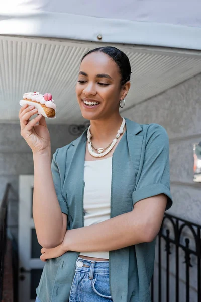 Pretty african american customer holding eclair with cream near sweet shop — Photo de stock