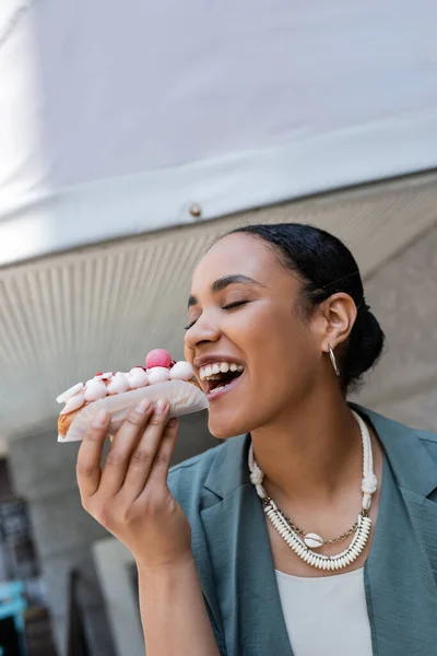 Young african american woman biting eclair near confectionery outdoors — Foto stock