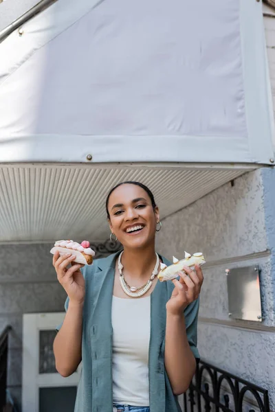 Happy african american woman holding tasty eclairs near sweet shop outdoors — Stock Photo