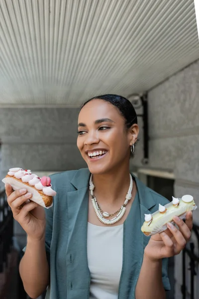 Cheerful young african american woman holding eclairs with cream near sweet shop — Fotografia de Stock