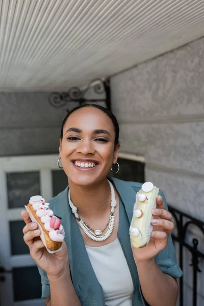 Positive african american woman holding delicious eclairs with cream near sweet shop outdoors — Fotografia de Stock