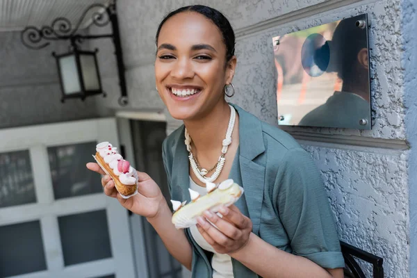 Positive african american customer holding sweet eclairs near confectionery outdoors — Photo de stock