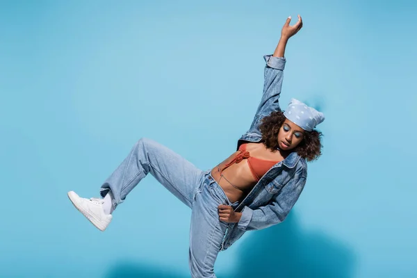 African american woman in denim clothes posing on one leg with arm up on blue background — Stockfoto