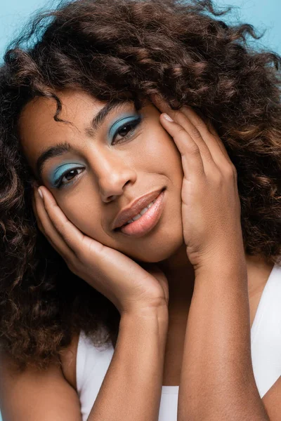Portrait of african american woman with makeup touching face and smiling at camera isolated on blue - foto de stock
