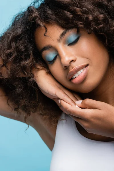 Happy african american woman with closed eyes and makeup holding hands near perfect face isolated on blue — Stock Photo