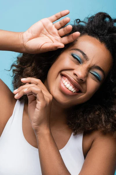 Portrait of cheerful african american woman with makeup laughing with closed eyes isolated on blue — Stock Photo