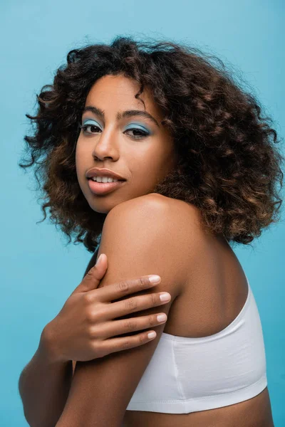 Brunette african american woman with perfect skin and makeup hugging her shoulder isolated on blue — стокове фото