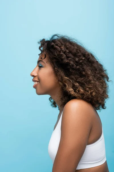 Вид збоку кучерявої і усміхненої афроамериканської жінки в білій верхній частині культури ізольовано на синьому — стокове фото