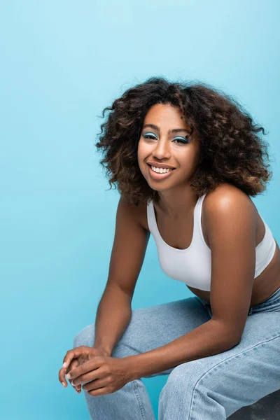 Brunette african american woman in white crop top smiling at camera while sitting isolated on blue — Stockfoto