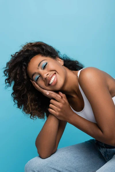 Cheerful african american woman with blue eye shadows holding hand near face and looking at camera isolated on blue — стоковое фото