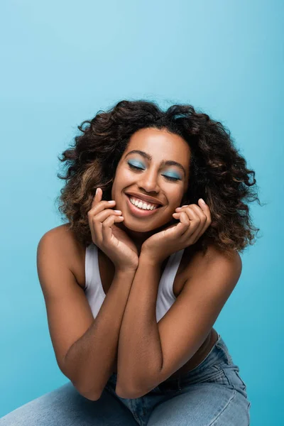 Pleased african american woman with curly hair and makeup holding hands near face isolated on blue — Stock Photo