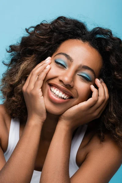 Joyful african american woman with perfect skin and makeup smiling at camera isolated on blue — стокове фото