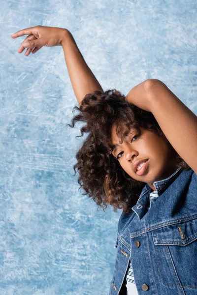 Curly african american woman in denim vest looking at camera while posing on blue background — Photo de stock