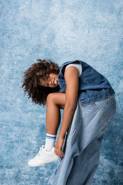 Модна афроамериканська жінка в рваних джинсах посміхається на камеру на синьому фоні — стокове фото