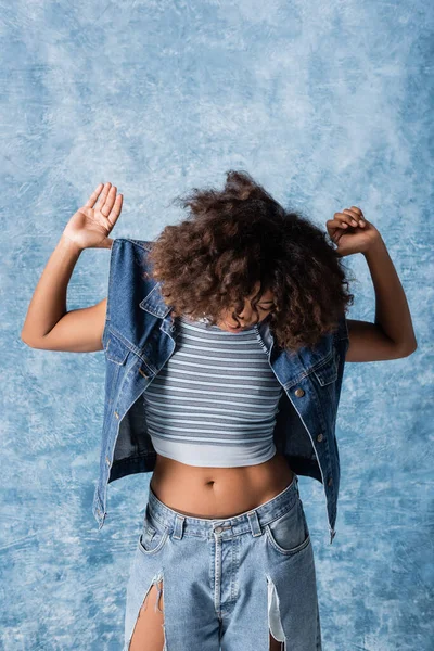 African american woman with face obscured by curly hair posing in denim vest on blue background — Stockfoto