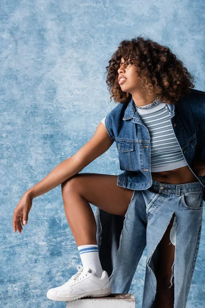 African american woman in stylish denim clothes stepping on stone block and looking away on blue background — Foto stock