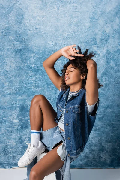 African american woman in stylish denim clothes with closed eyes and hands near head on blue background — Photo de stock