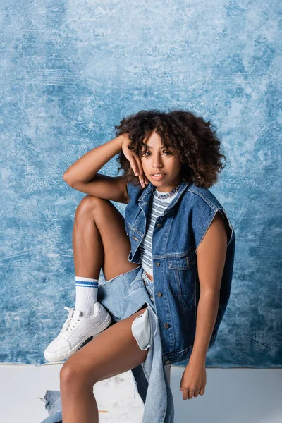 African american woman in fashionable denim clothes sitting and looking at camera on blue background — Fotografia de Stock