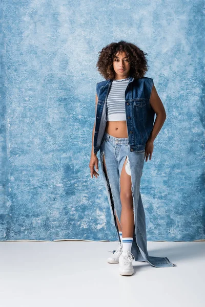 Full length of trendy african american woman in torn jeans and denim vest standing on blue background — Stockfoto