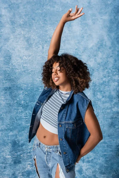 African american woman in denim vest and crop top standing with closed eyes and raised hand on blue background — Stockfoto