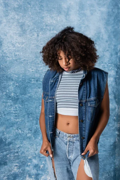 Brunette african american woman in striped crop top and denim vest on blue background — Foto stock