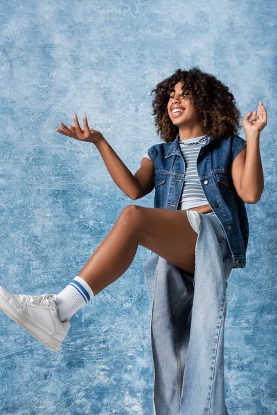 Щаслива афроамериканська жінка в рваних джинсах, стоячи на одній нозі і вказуючи рукою на синьому фоні — стокове фото