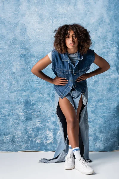 African american woman in ripped jeans and denim vest posing with hands on waist on blue background — стокове фото