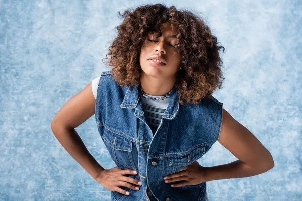 Stylish african american woman in denim vest standing with hands on waist on blue background — стоковое фото