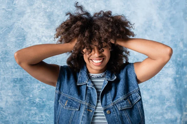 Cheerful african american woman in denim vest touching curly hair on blue background - foto de stock