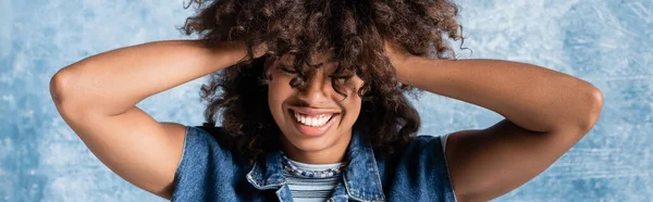 Smiling african american woman with closed eyes touching wavy hair on blue background, banner — Stockfoto