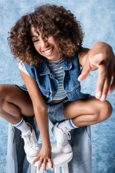 Happy african american woman in denim clothes pointing at camera while sitting on haunches on blue background — стоковое фото