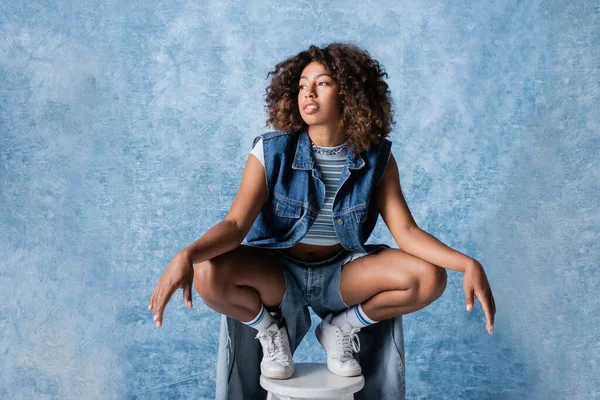 Full length of african american woman in trendy denim clothes posing on stool on blue background - foto de stock