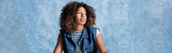 Curly african american woman in denim vest looking away on blue background, banner — Stockfoto