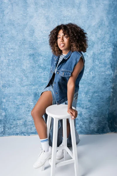 Full length of african american woman in white sneakers and denim clothing standing near stool on blue background — Stock Photo