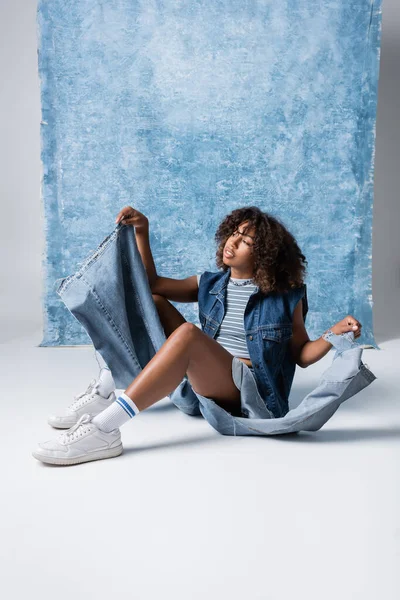 African american woman in torn jeans sitting on floor near blue drapery — Fotografia de Stock