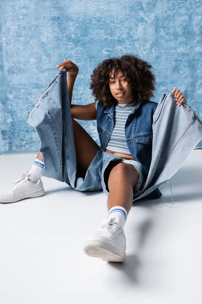 Fashionable african american woman sitting on floor in ripped jeans on blue background — Stock Photo