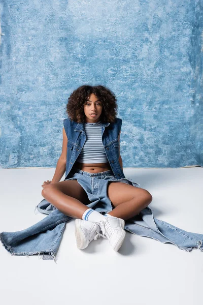 Full length of african american woman in ripped jeans and sneakers sitting on floor on blue background — Stockfoto