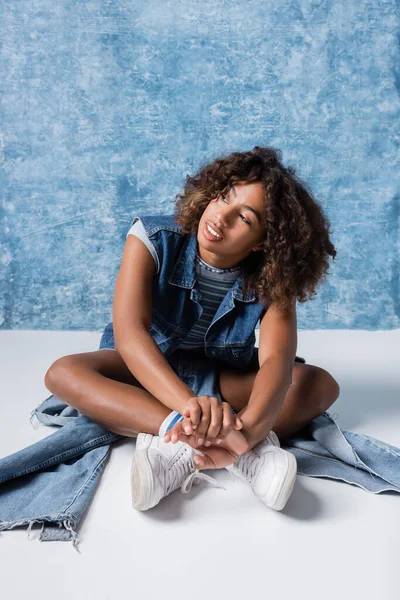 African american woman in white sneakers and denim clothes sitting with crossed legs on blue background — стоковое фото