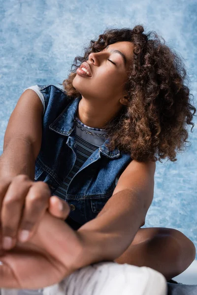 Sensual african american woman in denim vest posing with closed eyes on blue background — Foto stock