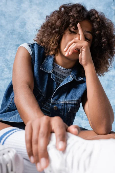 Brunette african american woman in denim vest obscuring face with hand on blue background - foto de stock