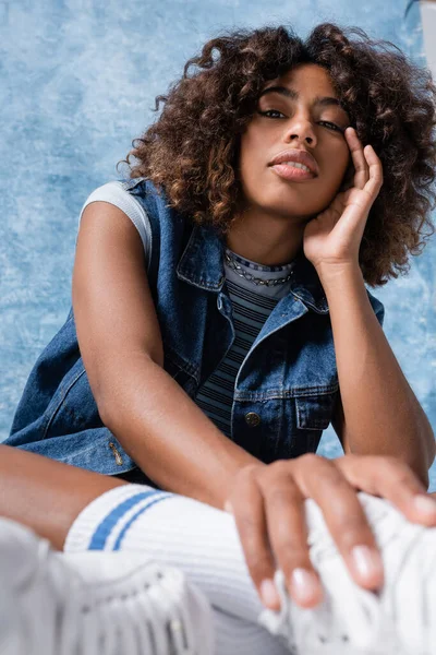 Trendy african american woman in denim vest holding hand near face and looking at camera on blue background — Fotografia de Stock