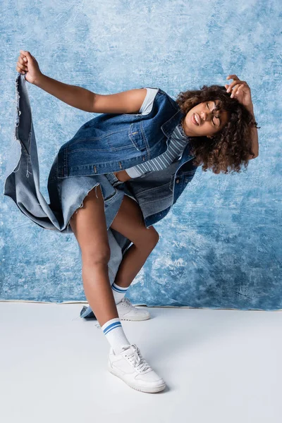 Full length of african american woman posing in torn jeans on blue background — Fotografia de Stock