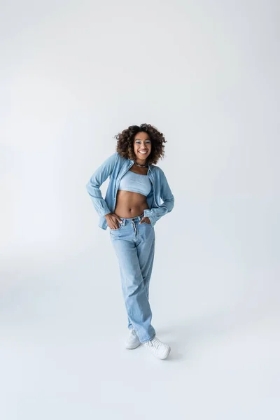 Full length of pleased african american woman standing with hands in pockets of blue jeans on grey background — Stockfoto
