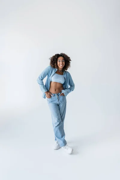 Full length of pleased african american woman standing with hands in pockets of blue jeans on grey background — Stock Photo