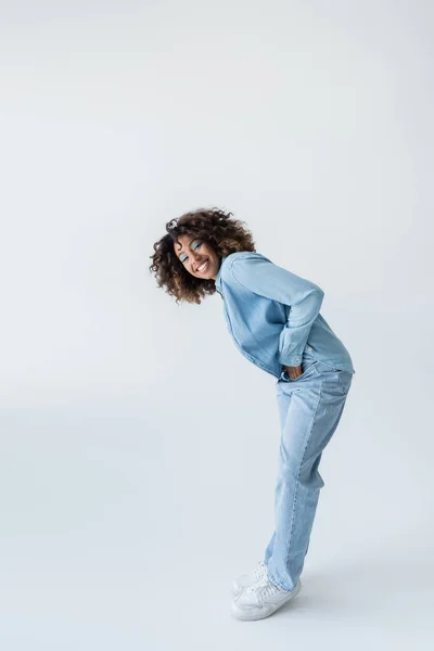 Full length of happy african american woman in denim clothes standing with hand in pocket on grey background — стоковое фото