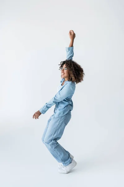 Side view of joyful african american woman posing in denim clothes with arm up on grey background — Stock Photo