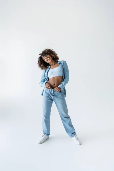 Full length of joyful african american woman in blue denim clothes and sneakers on grey background — Stockfoto