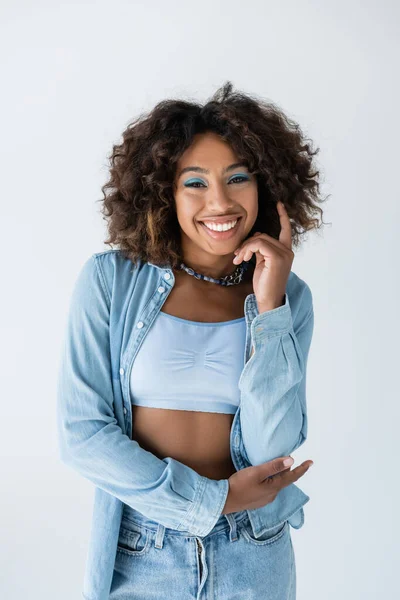 Positive african american woman in white crop top and denim shirt looking at camera isolated on grey - foto de stock