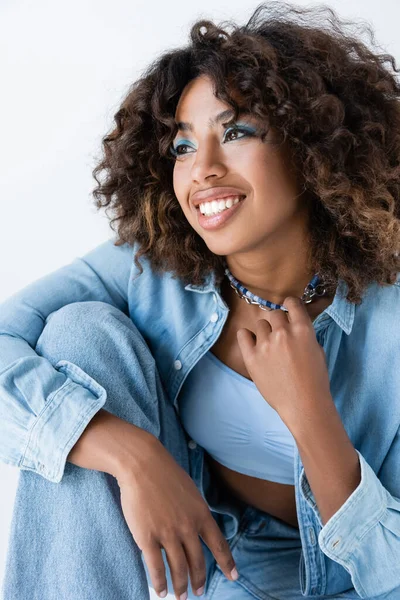Pleased african american woman in denim shirt touching necklaces and looking away isolated on grey — Stock Photo
